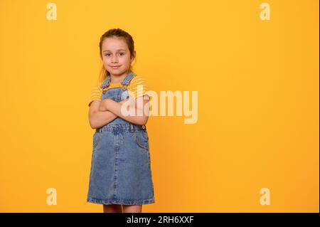 Portrait of a Caucasian beautiful charismatic emotional little girl 6 years old, wearing blue denim overalls, smiles looking at camera, posing with ar Stock Photo