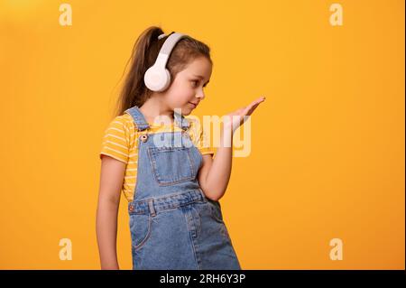 Caucasian adorable lovely cute little child girl wearing wireless headphones, dressed in yellow t-shirt and denim overalls, smiles looking at her hand Stock Photo