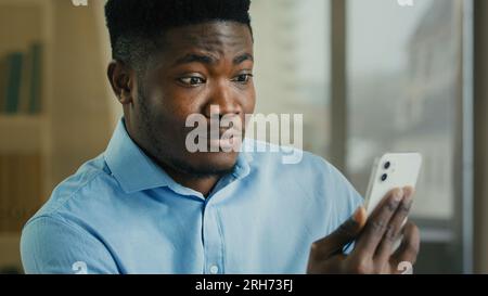 Surprised african businessman young office worker millennial happy amazed male man winner use mobile phone app celebrate good news victory win online Stock Photo