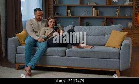 Diverse man and woman interracial couple homeowners relax on sofa talk video call communicate online with mobile phone web app happy diverse wife Stock Photo