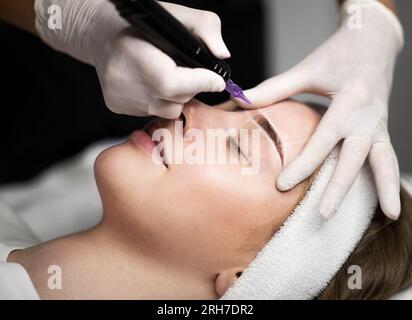 Cosmetologist applying permanent makeup on eyebrows of beautiful young female Stock Photo