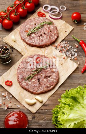 Raw burger patties with spices and rosemary on parchment, sauce, fresh vegetables, hamburger ingredients on a wooden background. Stock Photo