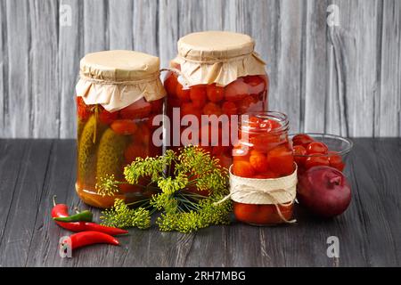 Canned cherry tomatoes and cucumbers in jars and a bowl, hot chili peppers, onions on a wooden background. Homemade preserves, pickled vegetables. Stock Photo