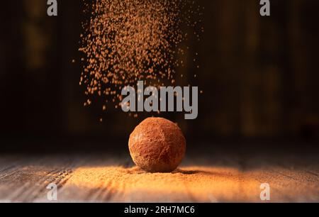 Cocoa powder is sprinkled on a chocolate truffle under a beautiful light. A ball of chocolate truffle is sprinkled with cocoa on a dark background Stock Photo