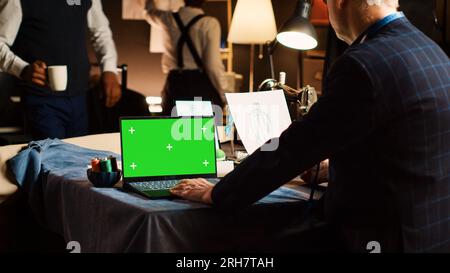 Old tailor using laptop with greenscreen, experienced fashion designer looking at chroma key template in atelier. Senior couturier designing clothes with isolated display in workshop. Stock Photo