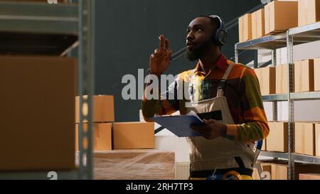 Young adult inspecting cargo logistics with inventory list, checking merchandise orders for retail delivery. Depot worker examining cardboard packages for distribution and stock production. Stock Photo