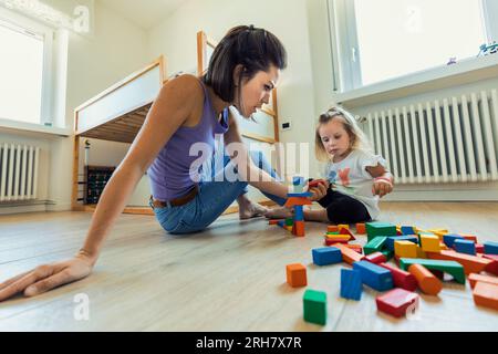 Mother and daughter engage in the playroom with wooden blocks, cups, developing skills, enjoying fun learning moments together. Stock Photo