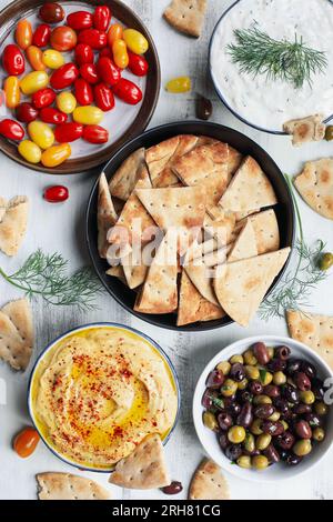 Mezze platter of pita bread surrounded by fresh tomatoes, olives, vegan tzatziki dip, and hummus over a white rustic table. Flatlay. Stock Photo