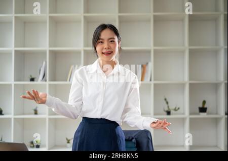 A beautiful and successful Asian businesswoman or female manager stands at her desk in her private office, smiling at the camera, and opens her palms. Stock Photo