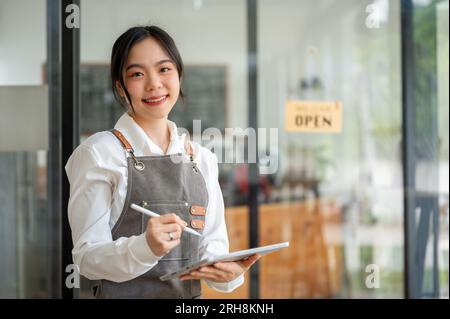 A friendly and pretty Asian female cafe worker or waitress in an apron stands in her cafe with a tablet in her hand. server, waitress, small business Stock Photo