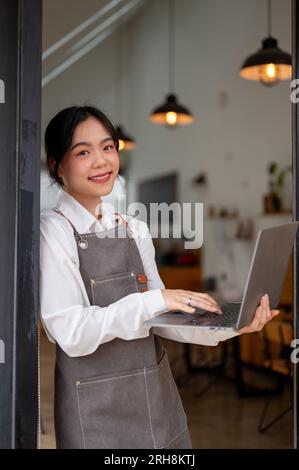 A portrait of a beautiful young Asian female coffee shop owner in an apron smiling at the camera and leaning on the wall of her coffee shop with a lap Stock Photo
