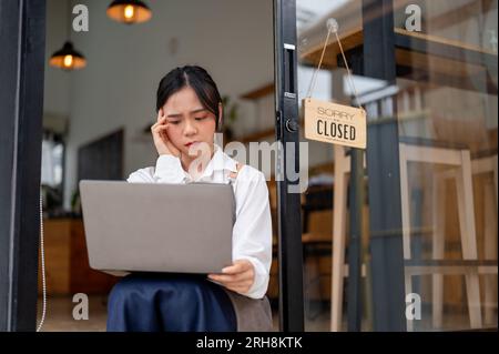 A stressed Asian female coffee shop or restaurant owner feels frustrated and overwhelmed with problems, looking at a laptop screen with a serious and Stock Photo
