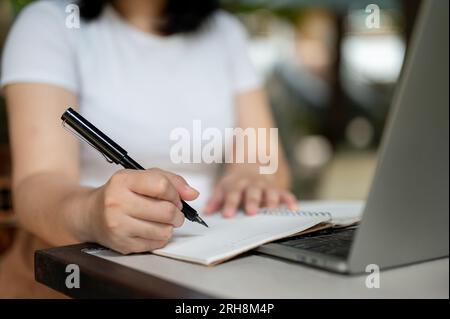 Cropped image of a woman working remotely at a coffee shop on the weekend, taking notes on a notebook, managing her work and using her laptop. Stock Photo