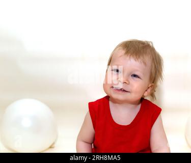 One-year-old toddler with blonde hair in red bodysuit sitting and smiling on white background with inflatable balloons. Wonderful kid sat down to rest Stock Photo