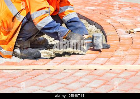 Bricklayer in old overalls squatting laying orange paving slabs on a sunny day. Copy space. Stock Photo