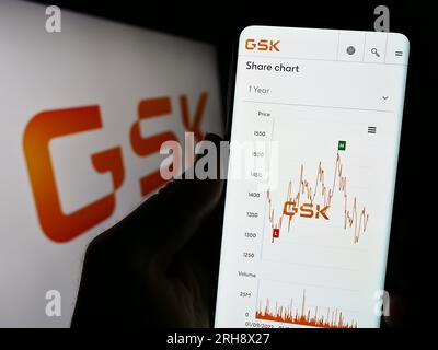 Person holding cellphone with webpage of British pharmaceutical company GSK plc on screen in front of logo. Focus on center of phone display. Stock Photo