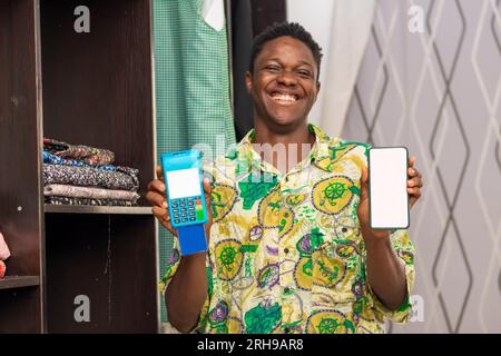 photo of a cheerful black businessman smiling while holding pos terminal and smartphone with empty white blank screen Stock Photo