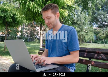 Young cheerful man freelancer working online using laptop while sitting on the bench outdoor. Remote job using wireless technology Stock Photo