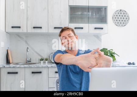 Young cheerful man stretching hands while working on laptop online at home and having break. Male freelancer using computer for distance work enjoying Stock Photo