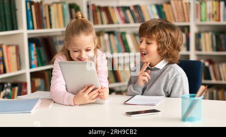 Two primary school kids sitting together at desk in classroom or library interior, using digital tablet computer Stock Photo