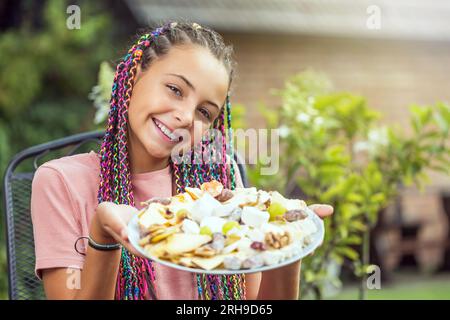 Little smiling long haired girl is holding a plate full of cheeses outdoors.  Horizontally. Stock Photo