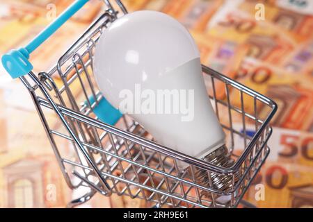 Electric energy saving bulb in a mini shopping cart on a money euro background. Electrical costs concept Stock Photo