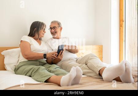 Smiling senior european man and woman watch video on phone, sit on bed in bedroom interior Stock Photo
