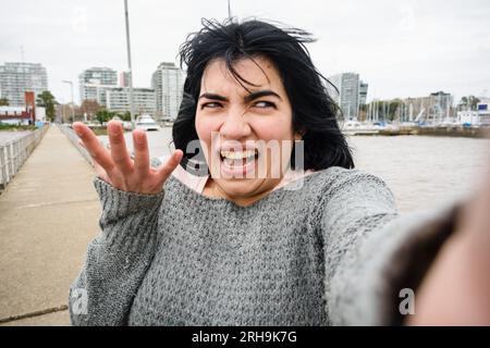 young venezuelan latin woman, standing on pier over river in buenos aires is laughing making funny selfie portrait with her phone, technology concept, Stock Photo