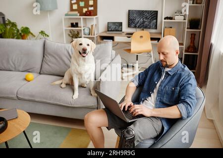 Portrait of adult man with disability using laptop in living room with white dog in background, copy space Stock Photo