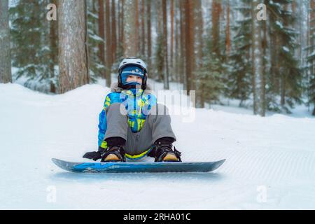 little boy sitting on snow putting his feet in snowboard bindings adjusting straps. Image with selective focus. High quality photo Stock Photo