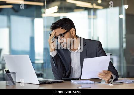 Serious thinking businessman at workplace, hispanic man upset thinking about financial reports, boss in business suit holding contract accounts, man unhappy with financial results of achievement. Stock Photo