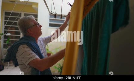 Elderly man doing domestic household chores in home backyard, removing dried clothes from drying hanger. candid everyday lifestyle scene in old age Stock Photo