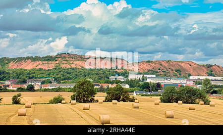 Edinburgh, Scotland, UK. 15th  August, 2023. UK Weather: Harvest at Greendykes shale bing sees yellow fields of baled hay before the cliffs of the waste product of the shale oil industry under turbulent sunshine sky.  Credit Gerard Ferry/Alamy Live News Stock Photo