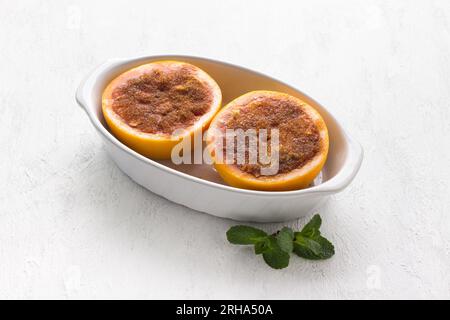 White ceramic baking dish with red grapefruit halves smeared with butter and sprinkled with sugar and cinnamon on a light gray table. Cooking step, co Stock Photo