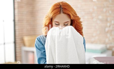 Young redhead woman smelling clean towel at laundry room Stock Photo