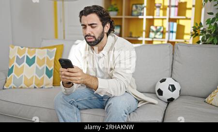 Young hispanic man watching soccer match using smartphone at home Stock Photo