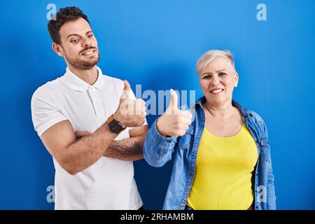 Young brazilian mother and son standing over blue background looking proud, smiling doing thumbs up gesture to the side Stock Photo