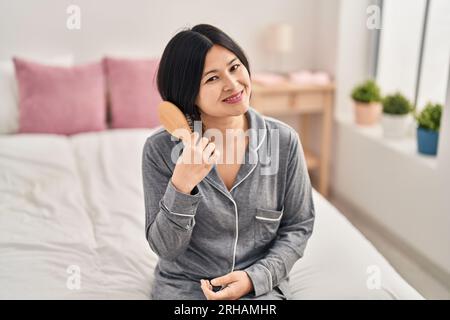 Young chinese woman combing hair sitting on bed at bedroom Stock Photo
