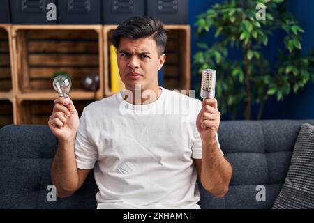 Hispanic man holding led lightbulb and incandescent bulb skeptic and nervous, frowning upset because of problem. negative person. Stock Photo