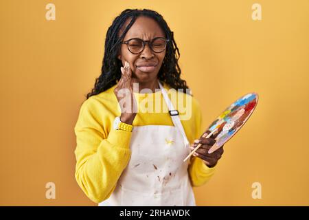 African woman holding painter palette touching mouth with hand with painful expression because of toothache or dental illness on teeth. dentist concep Stock Photo