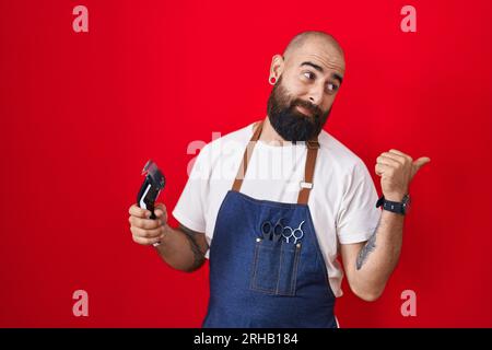 Young hispanic man with beard and tattoos wearing barber apron holding razor smiling with happy face looking and pointing to the side with thumb up. Stock Photo