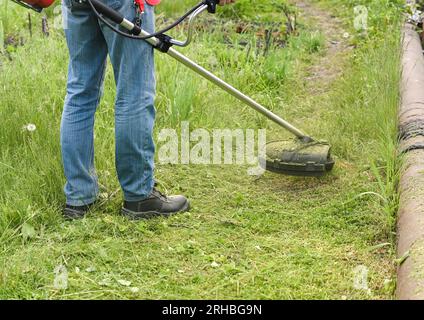 man mowing tall grass with electric or petrol lawn trimmer in city park or backyard. Process of lawn trimming with hand mower. Stock Photo