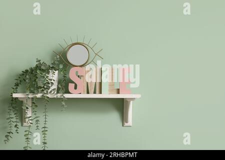 Shelf with small mirror, wooden letters spelling word SMILE and houseplant hanging on green wall Stock Photo