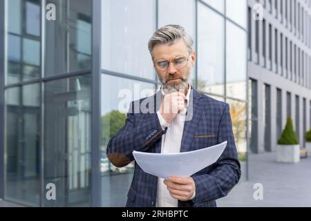 Serious thinking businessman boss with documents contracts invoices outside office building, mature experienced man in business suit looking at papers, with graphs financial reports. Stock Photo