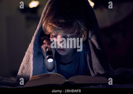 Focused boy reading book with interest and shining flashlight while lying on bed under warm blanket in dark room of apartment Stock Photo