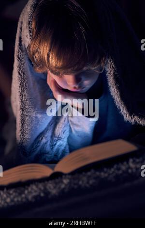 Focused boy wrapped in warm blanket reading on hand and reading interesting book while lying on bed in dark room Stock Photo