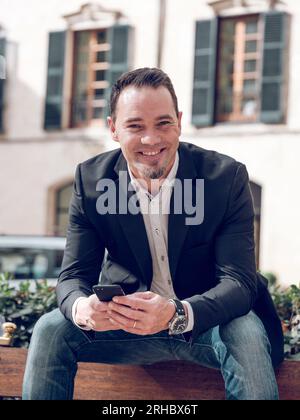 Delighted adult male manager with dark hair and bear in stylish smart casual outfit smiling happily and looking at camera while using mobile phone sit Stock Photo