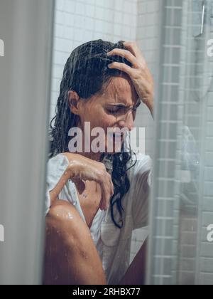 Dressed long haired lady crying in shower cabin while sitting under spraying water and touching forehead Stock Photo