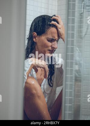 Sorrowing woman with long hair getting shower in white shirt while sitting behind transparent curtain Stock Photo