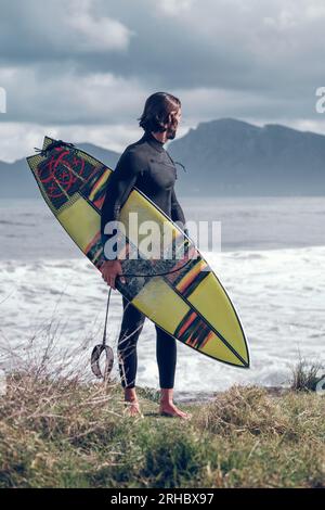 Full body of young muscular bearded male athlete in black wetsuit with surfboard in hand standing on grassy coast and looking away after practicing su Stock Photo